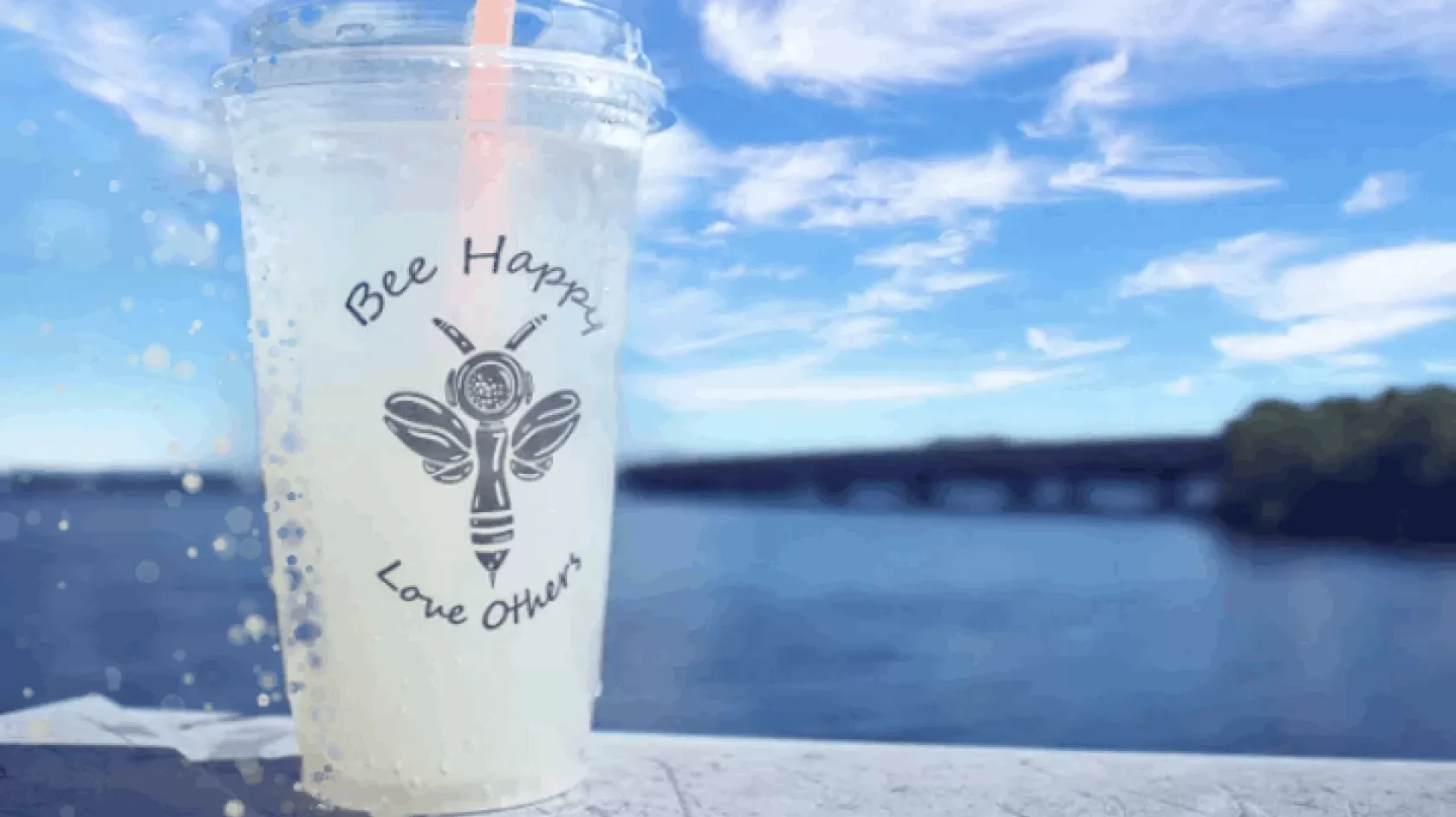 A cool beverage with a pink straw sit on the edge of a dock on the water